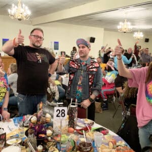 a group of young people at a trivia night standing around a table covered in snacks and gifts and raising the thumbs up hand sign